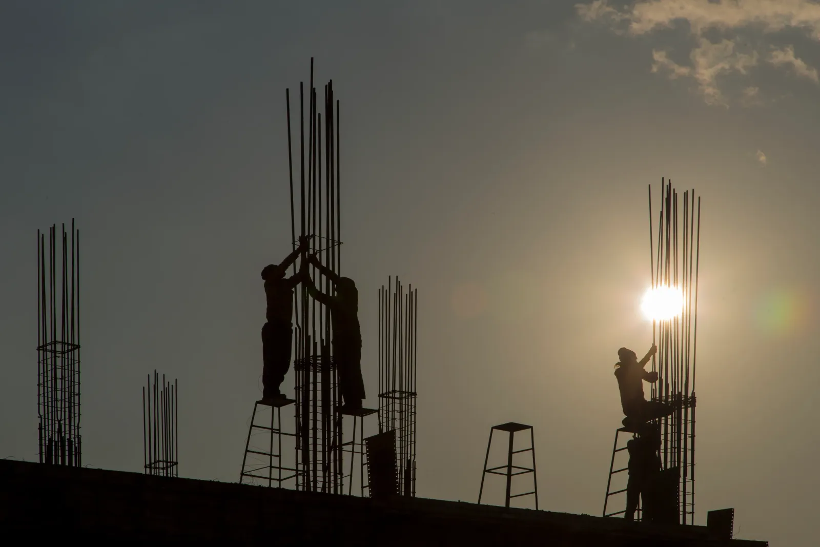 Operarios a contraluz sobre escaleras de tijera, atando las armaduras de espera de unos pilares, sin protección de borde.